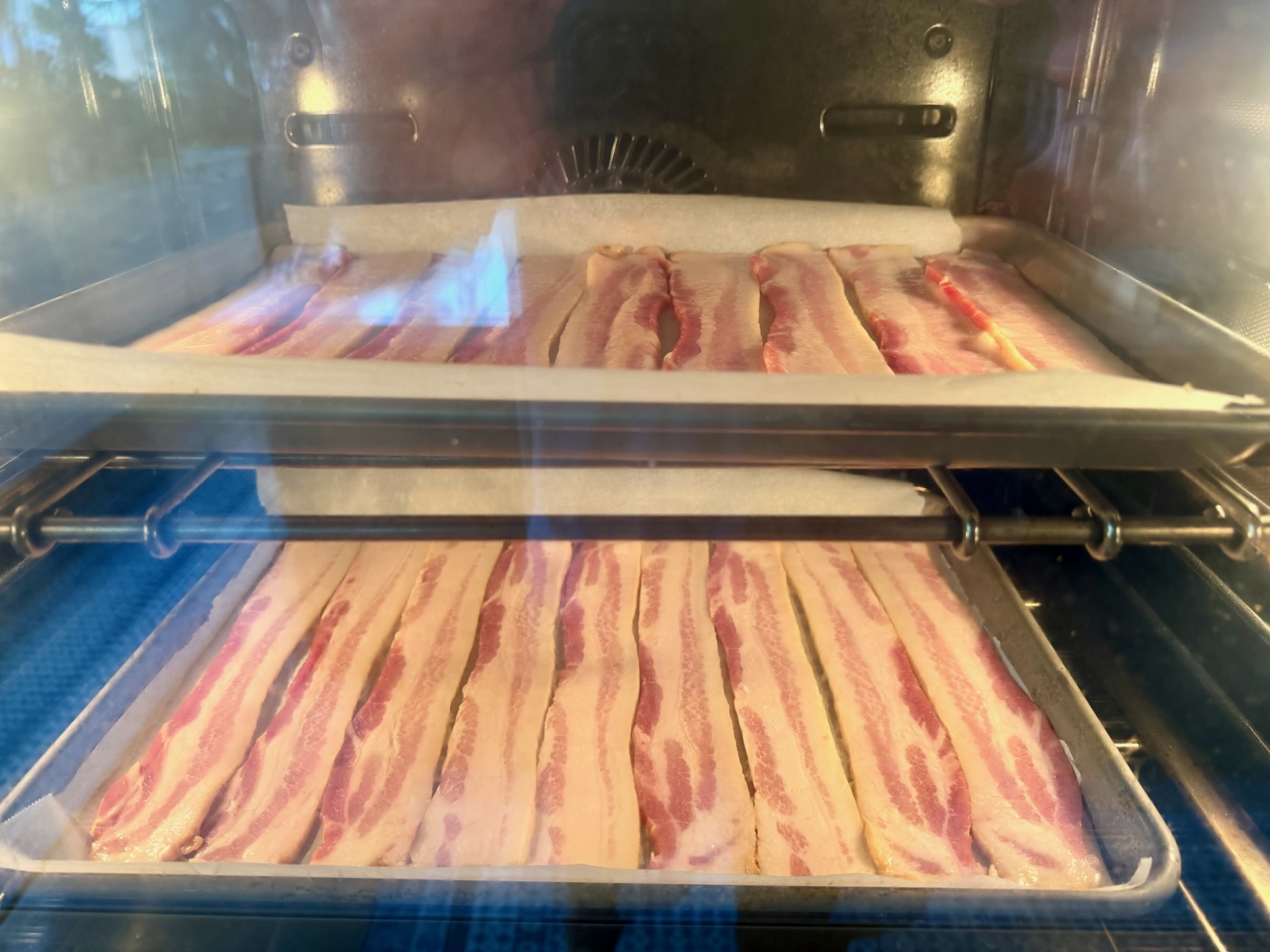 Thick-cut bacon on two parchment-lined sheet trays viewed through the oven door.
