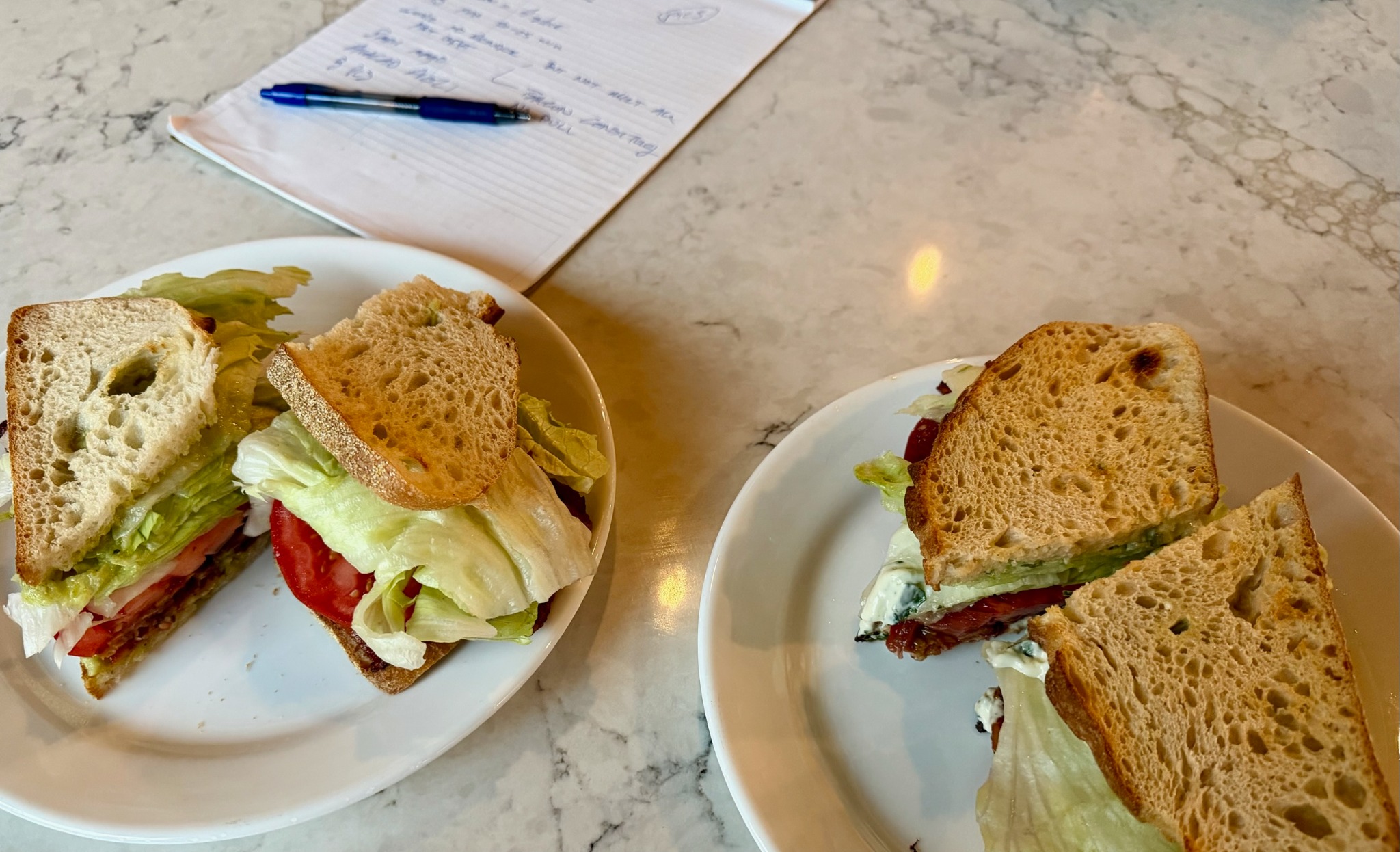 Two BLT test sandwiches on white plates beside handwritten notes and a blue pen.